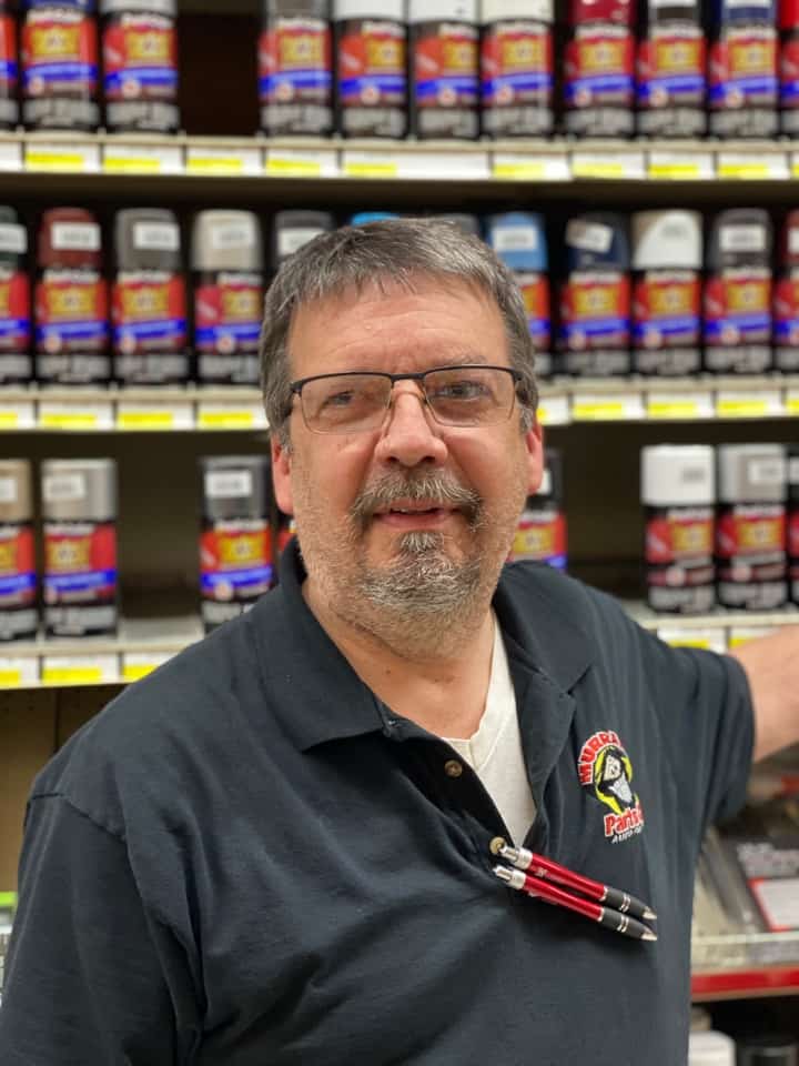 Middle age man with mustache and beard wearing glasses and black polo shirt in front of merchandise shelving.